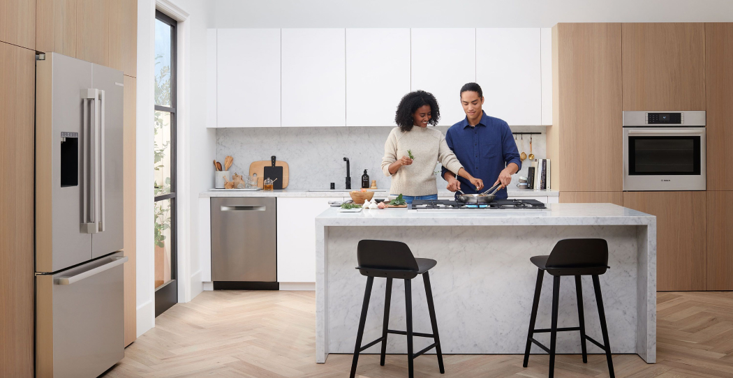 a photo of a couple cooking at a cooktop in the middle of a primarily white kitchen with some white wooden accents